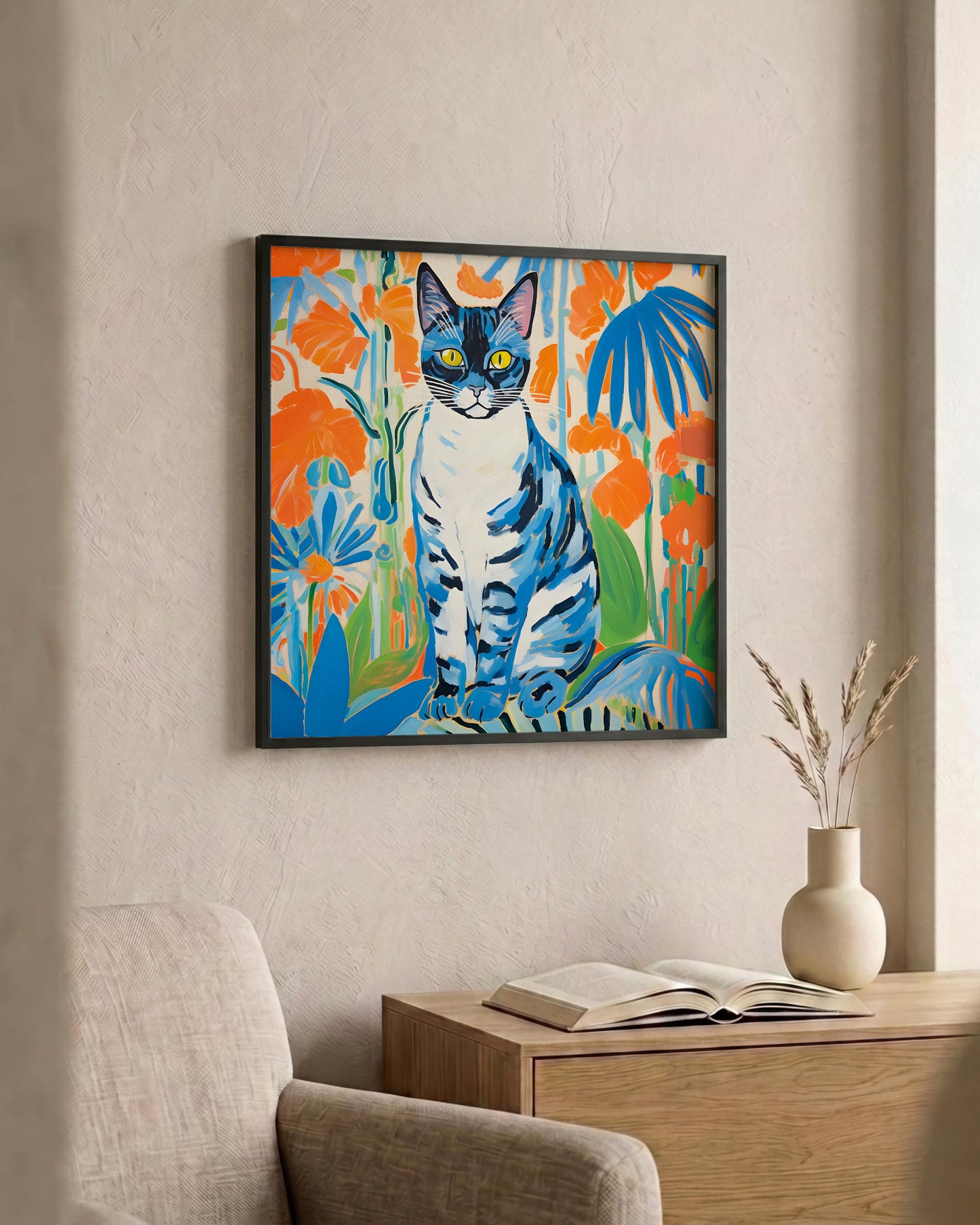 A framed square wall art print showing a colorful cat surrounded by bright flowers, displayed on a beige wall above a wooden side table with an open book and a small vase.