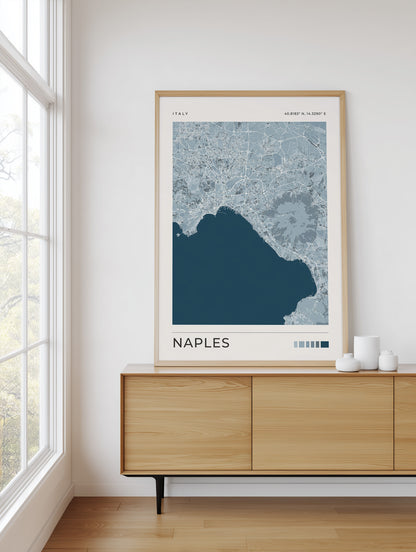 A framed Naples city map art print with a light wood frame, displayed on a light wood sideboard in a bright, modern room next to a window.