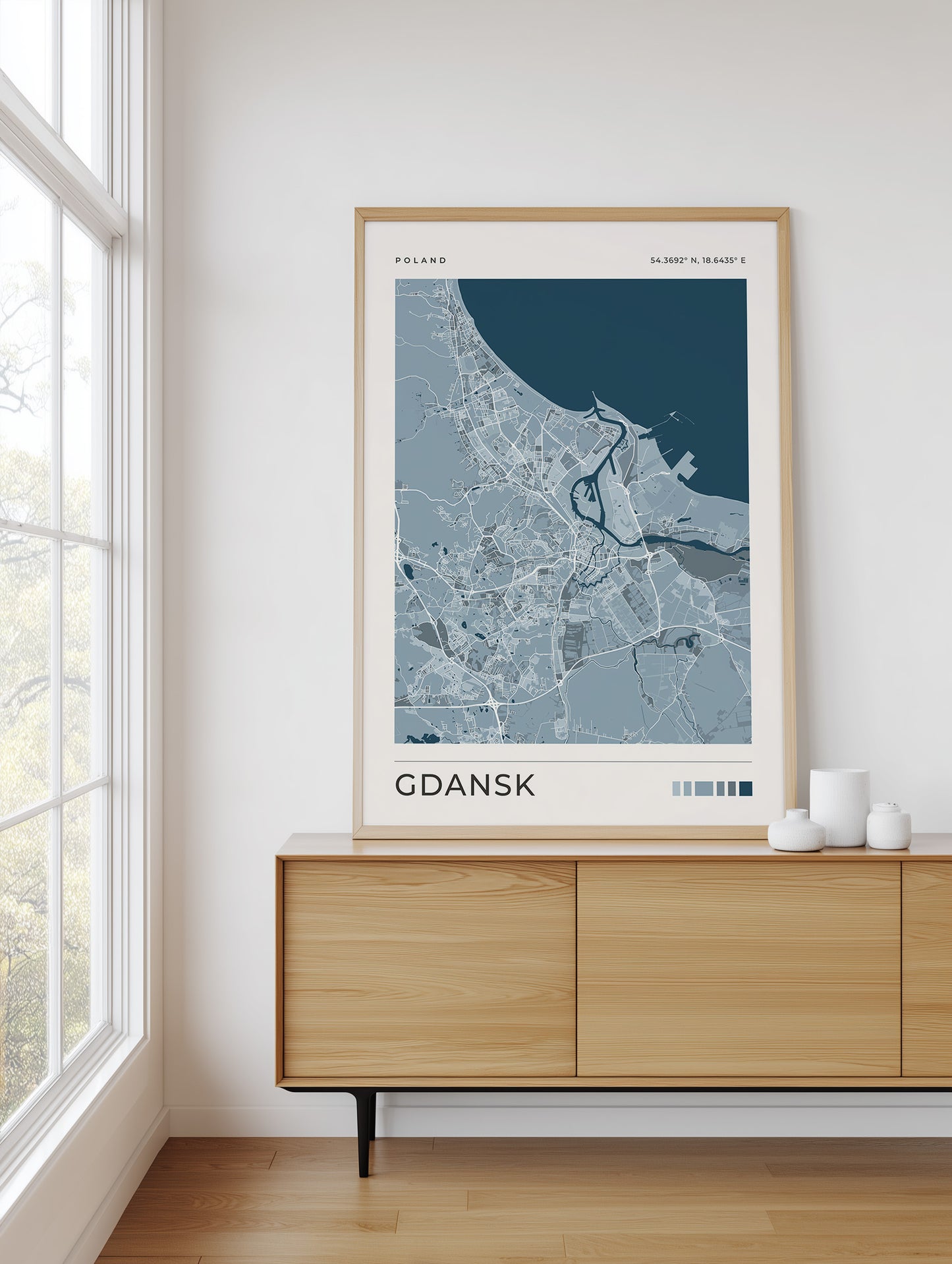 A framed street map art print of Gdansk, Poland, displayed on a light wood sideboard in a bright, modern room next to a window.