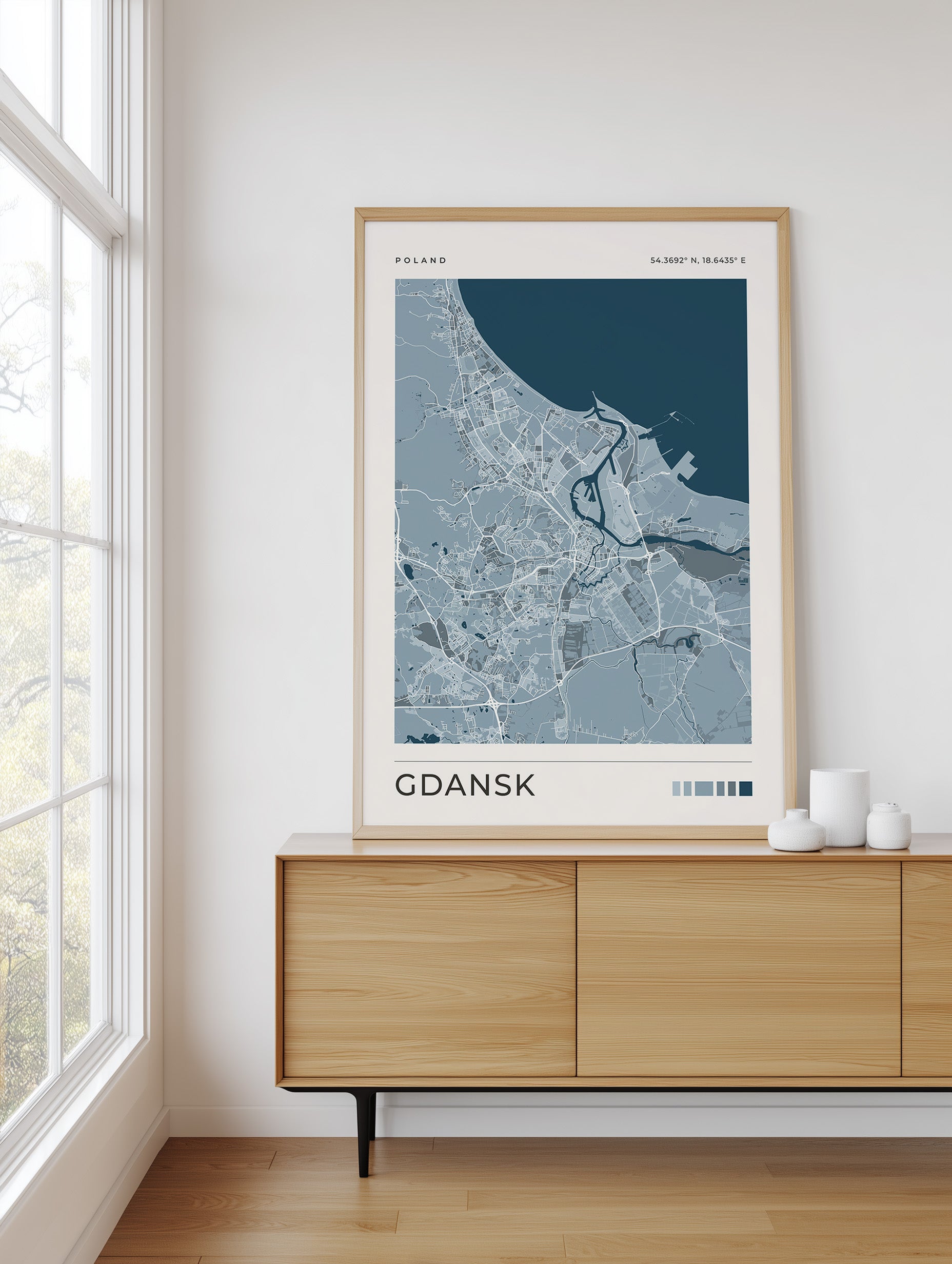A framed street map art print of Gdansk, Poland, displayed on a light wood sideboard in a bright, modern room next to a window.