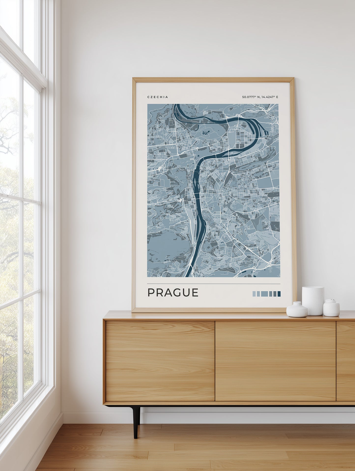A framed art print of a Prague city map, featuring a light blue and gray map design with a dark blue river, displayed on a light wood sideboard in a bright room next to a window.