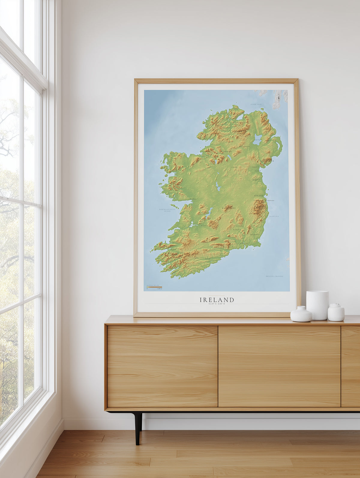 A framed topographical map of Ireland, featuring green landmasses and a light blue sea, displayed on a light wood sideboard in a bright room next to a window.