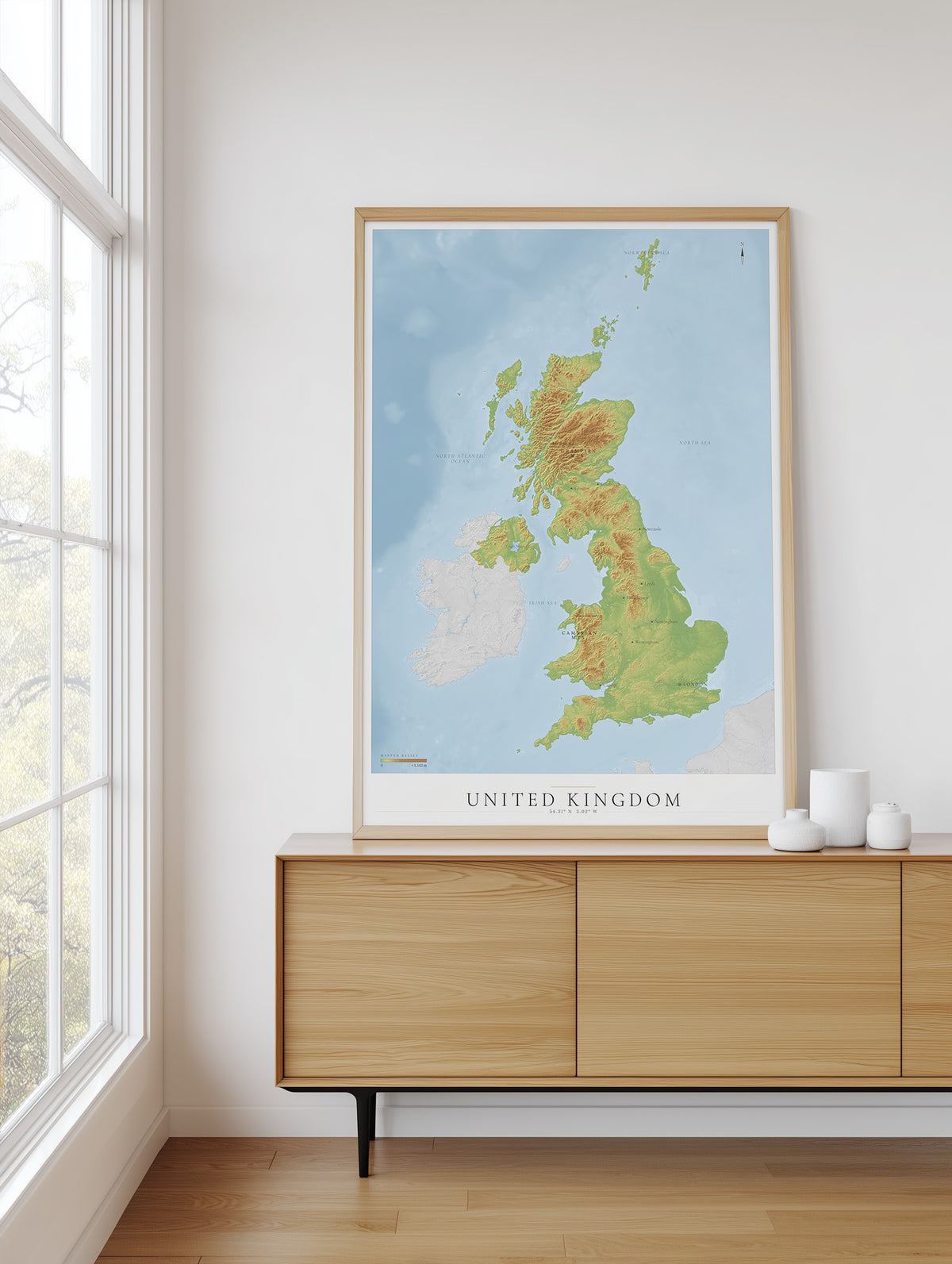A framed topographical map poster of the United Kingdom, featuring green and yellow landmasses on a light blue ocean background, styled on a light wood sideboard in a bright room next to a window.