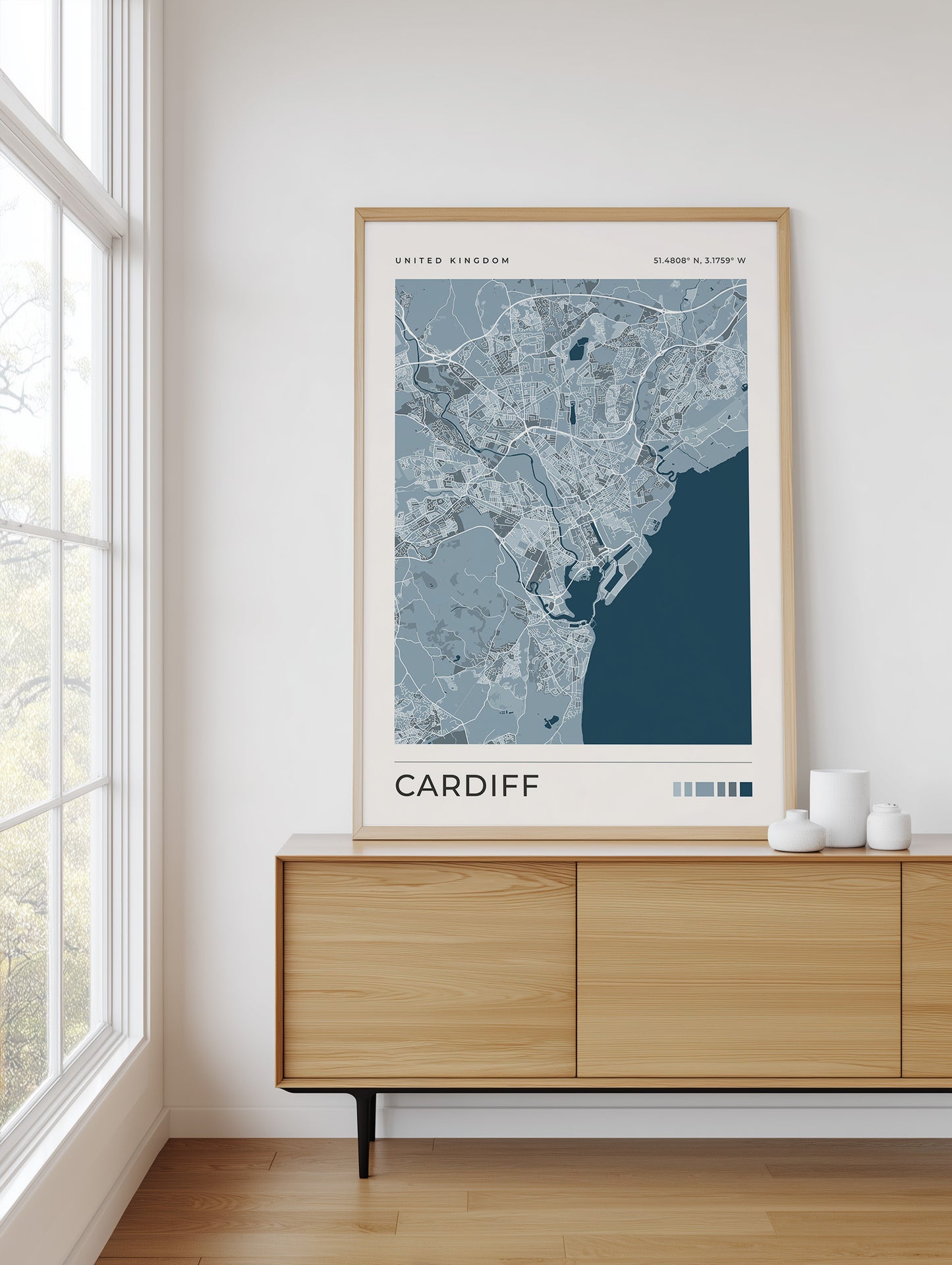 A framed city map print of Cardiff, United Kingdom, styled on a light wood sideboard in a bright, modern room next to a window.