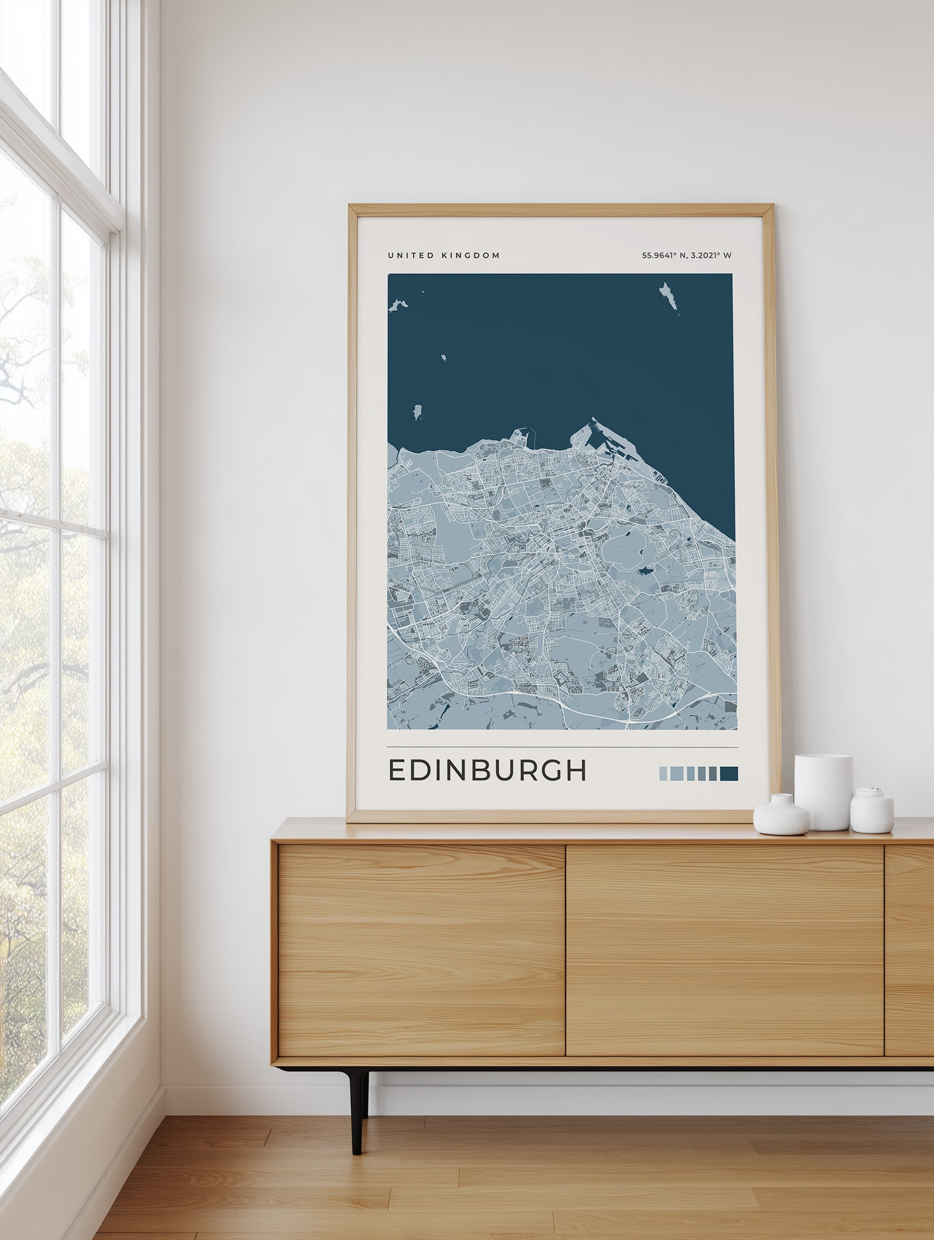 A framed Edinburgh city map poster with a light wood frame, displayed on a wooden sideboard in a bright room next to a window.