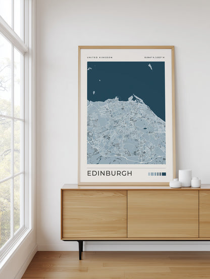 A framed Edinburgh city map poster with a light wood frame, displayed on a wooden sideboard in a bright room next to a window.