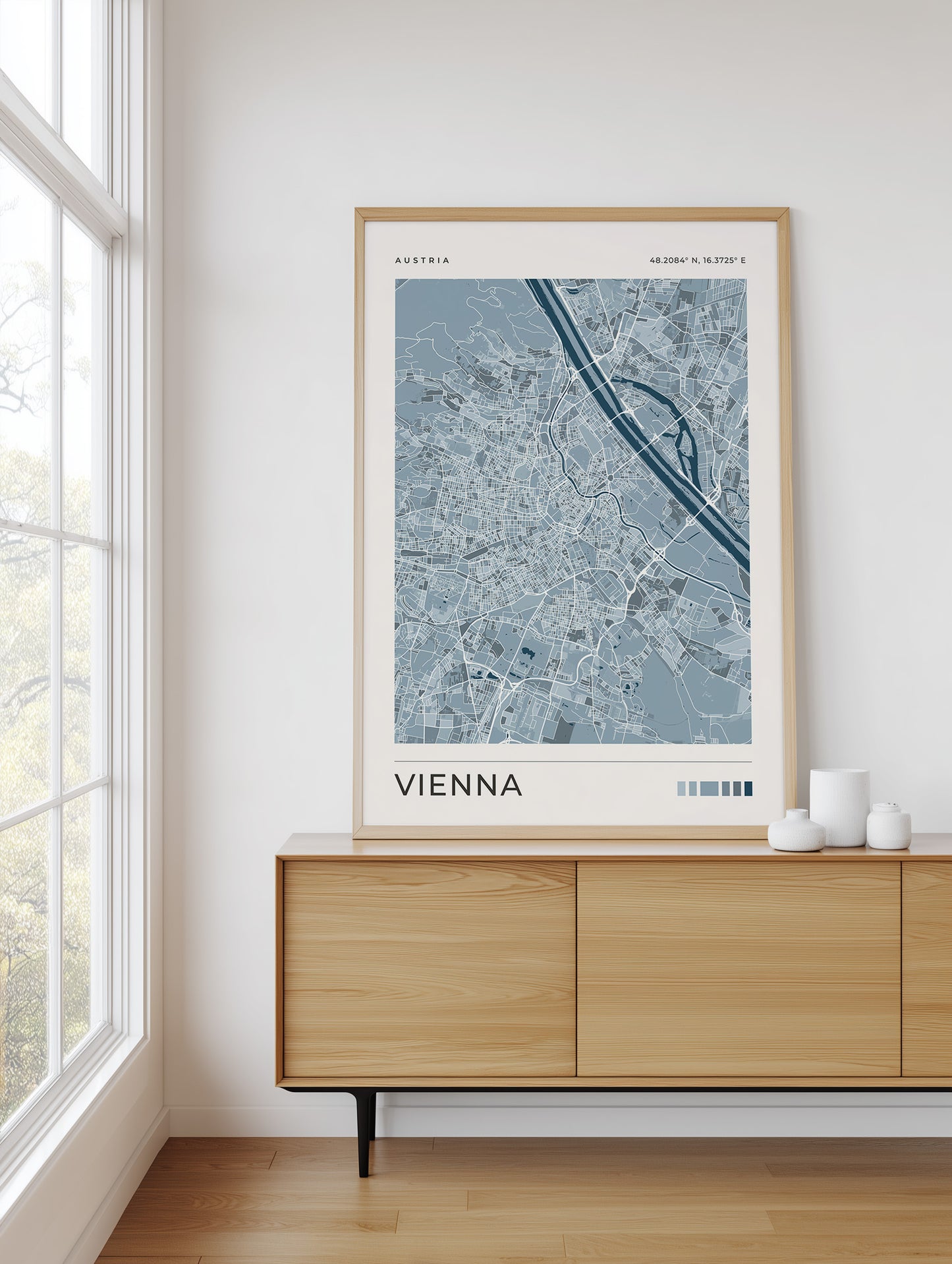 A framed art print of a Vienna street map in blue and gray tones, styled on a light wood sideboard in a bright, modern room next to a window.