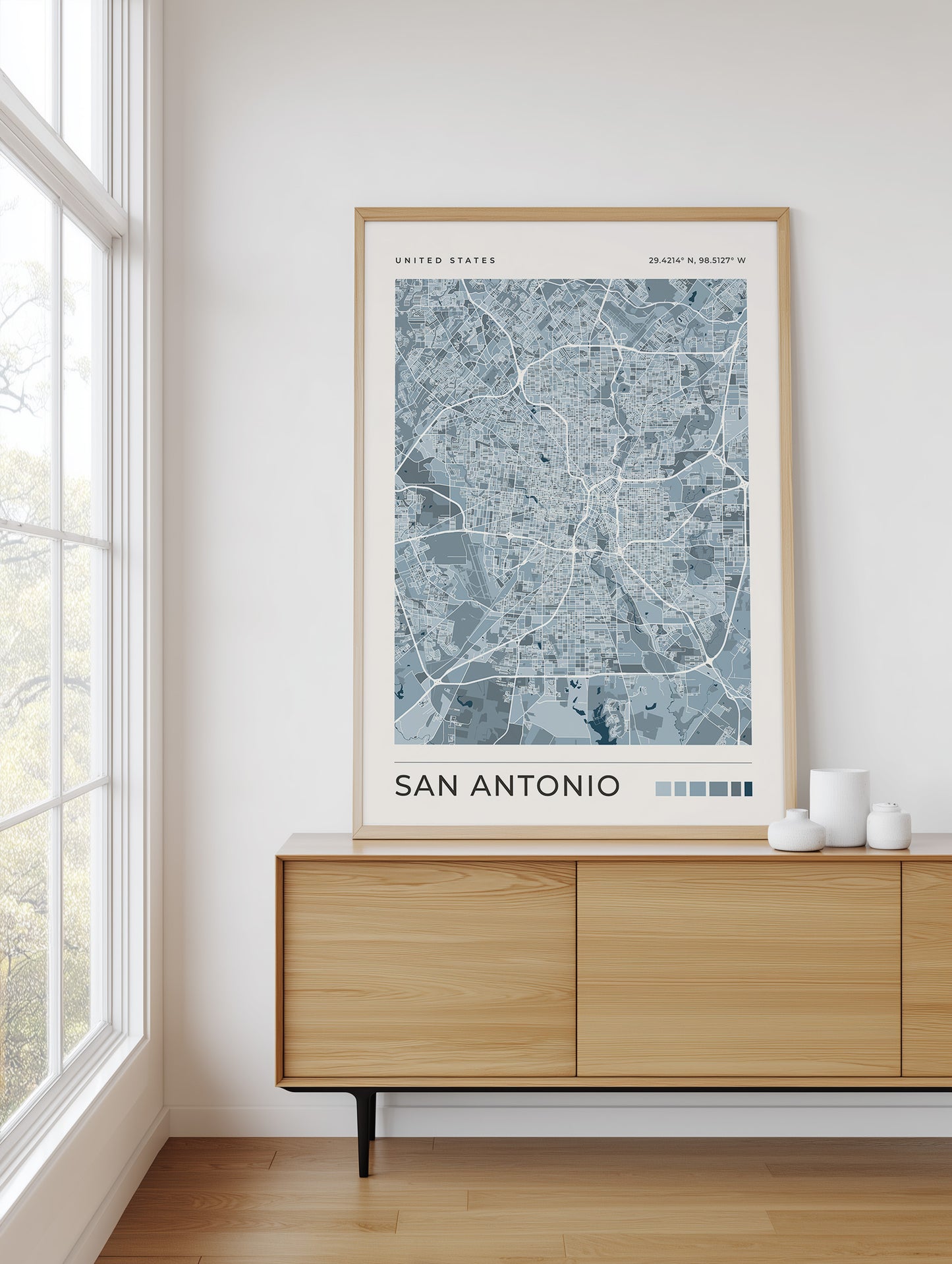 A framed San Antonio city map print hangs on a white wall above a light wood sideboard in a bright room with a window and small white decorative vases.