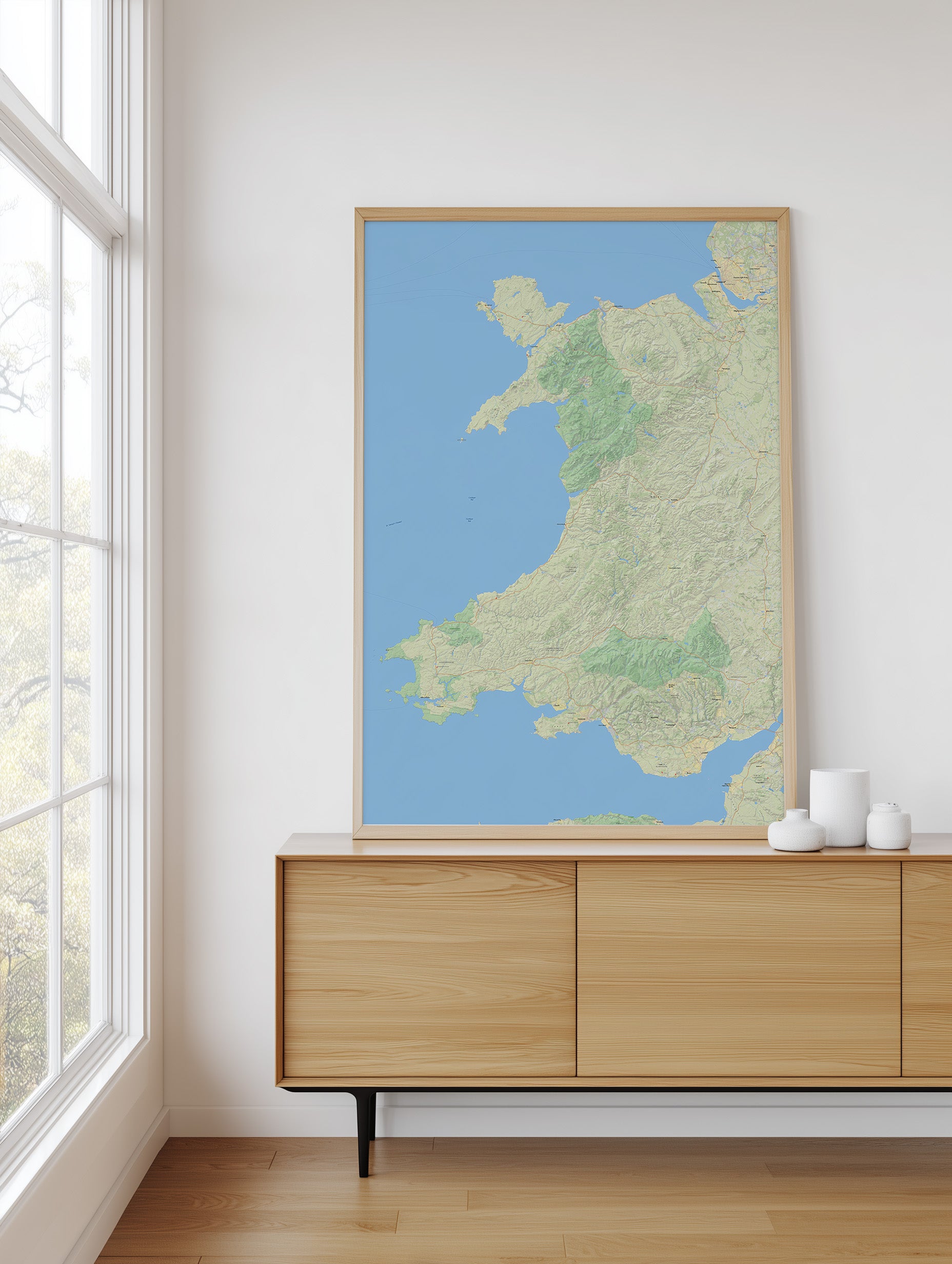 A framed topographical map print of Wales, featuring light green land and a blue sea, displayed on a light wood sideboard in a bright, modern room next to a window.