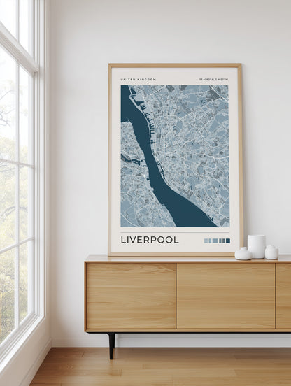 A framed art print of a blue and white map of Liverpool, United Kingdom, displayed on a light wood sideboard in a bright room with a window.