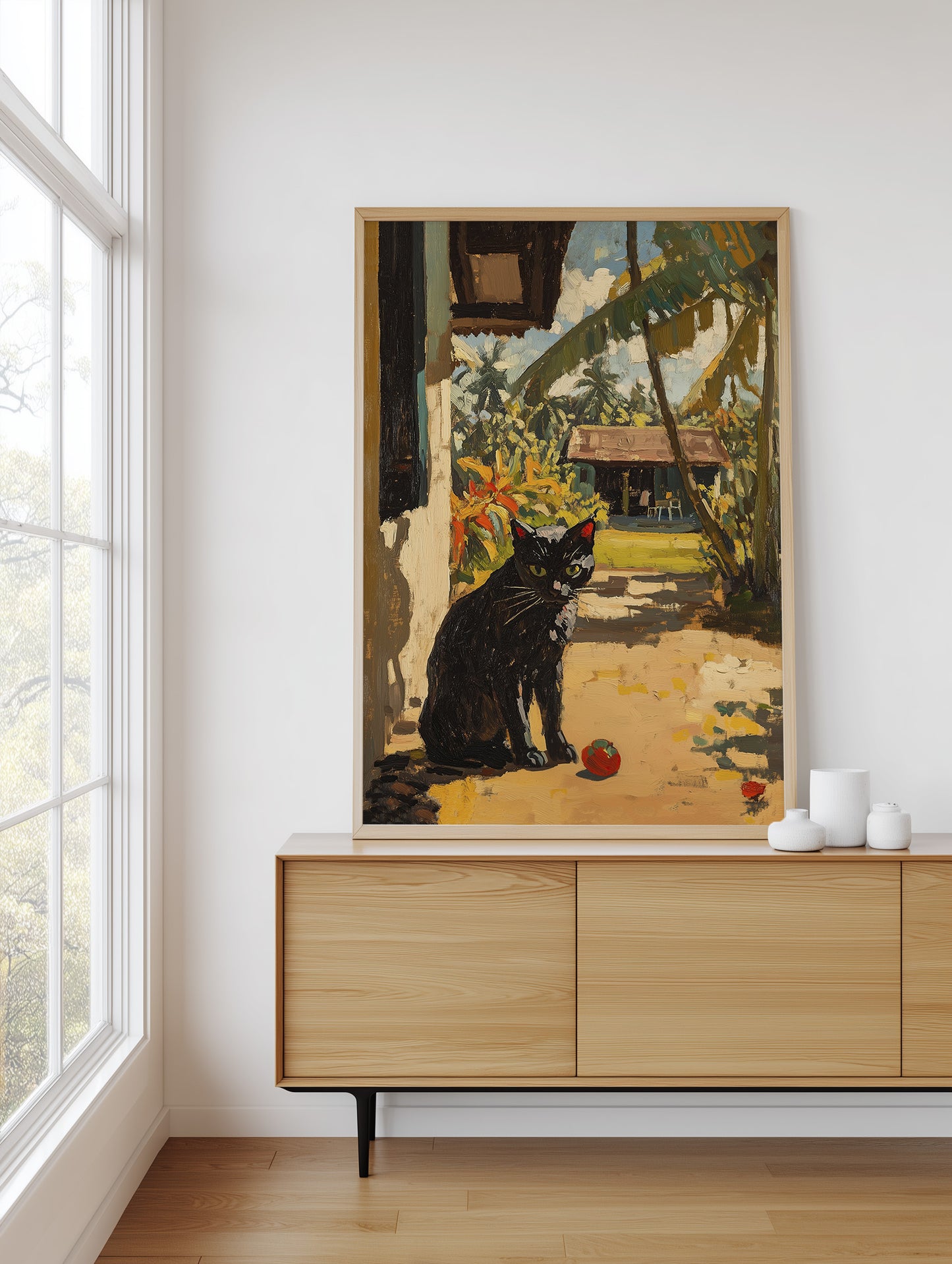 A framed art print depicting a black cat in a tropical outdoor setting, displayed on a light wood console table in a modern room next to a window.