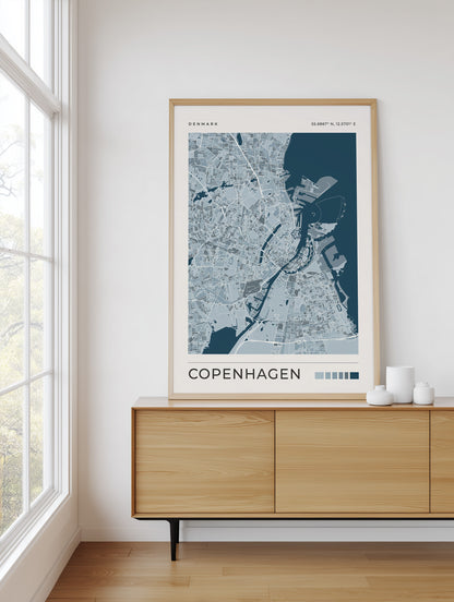 A framed Copenhagen city map poster in navy blue and light blue tones, styled on a light wood sideboard in a bright room next to a window.