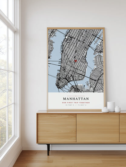 A framed Manhattan city map art print hangs on a white wall above a light wood sideboard in a bright room with a window and small white decorative vases.