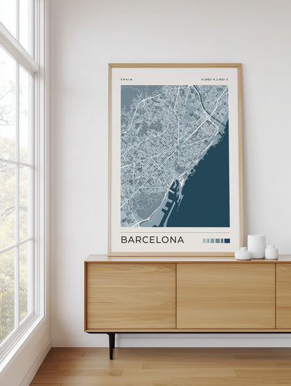 A framed Barcelona city map poster with a light wood frame, displayed on a light wood sideboard in a bright room next to a window.