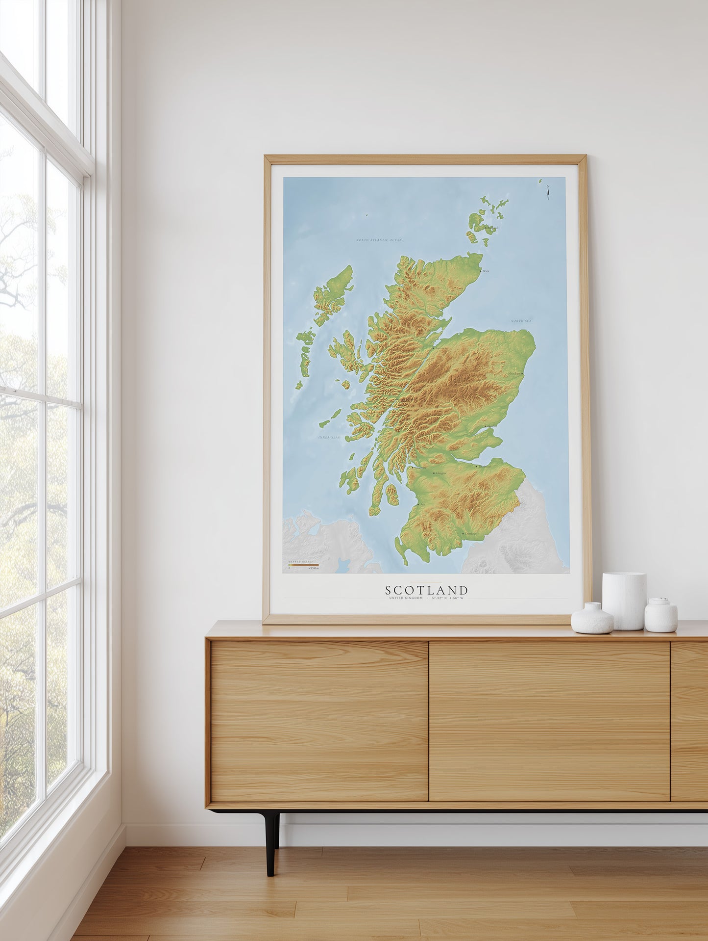A framed topographic map poster of Scotland, featuring green and yellow landmasses on a light blue background, styled on a light wood sideboard in a bright room next to a window.
