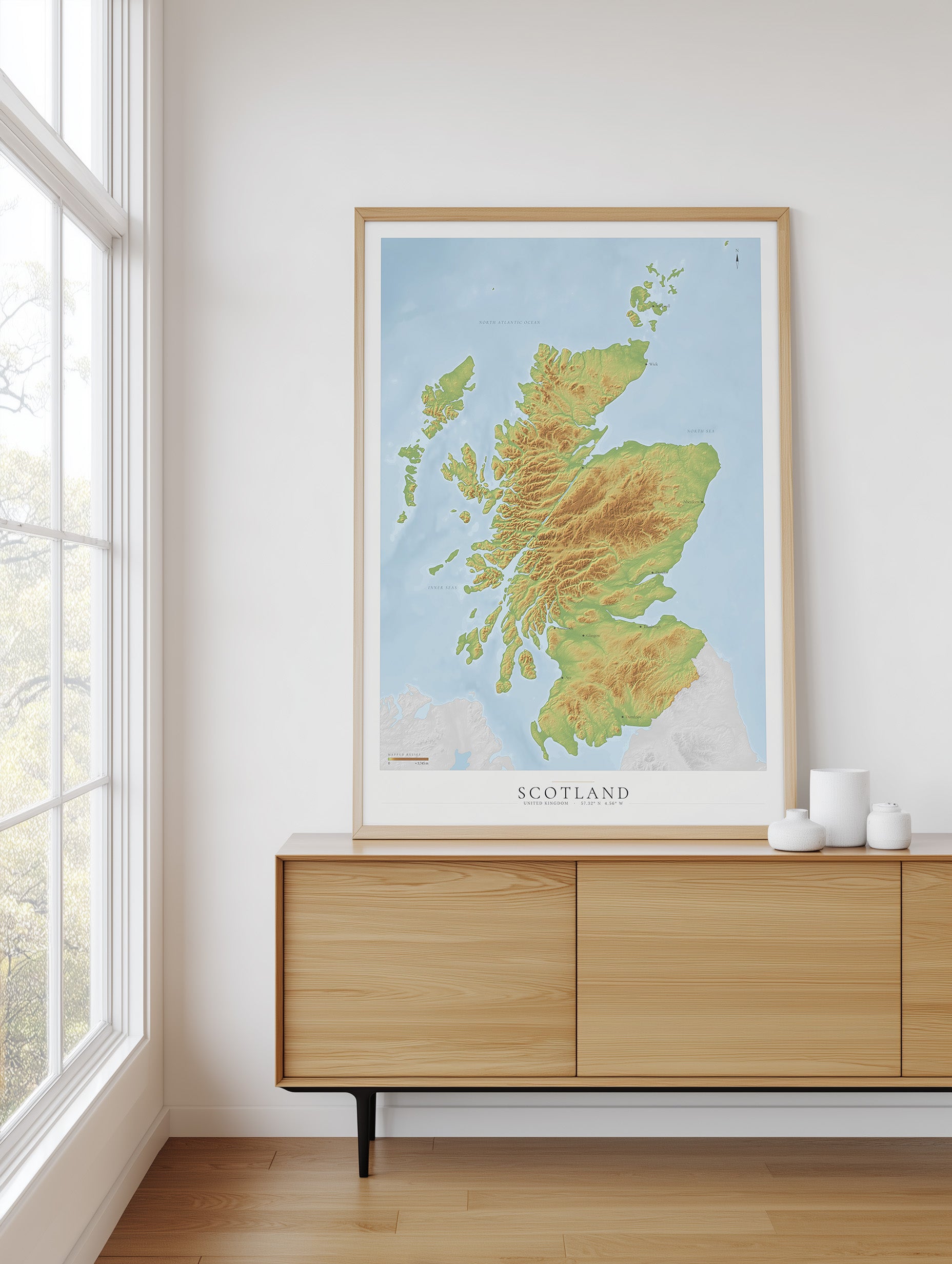 A framed topographic map poster of Scotland, featuring green and yellow landmasses on a light blue background, styled on a light wood sideboard in a bright room next to a window.
