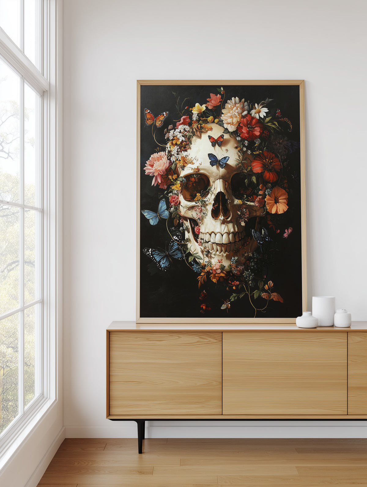 A framed art print featuring a detailed skull surrounded by colorful flowers and butterflies on a dark background, displayed on a light wood sideboard in a bright room next to a window.