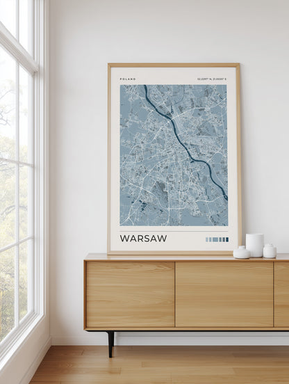 A framed art print of a light blue and white street map of Warsaw, Poland, styled on a light wood sideboard in a bright room next to a window.