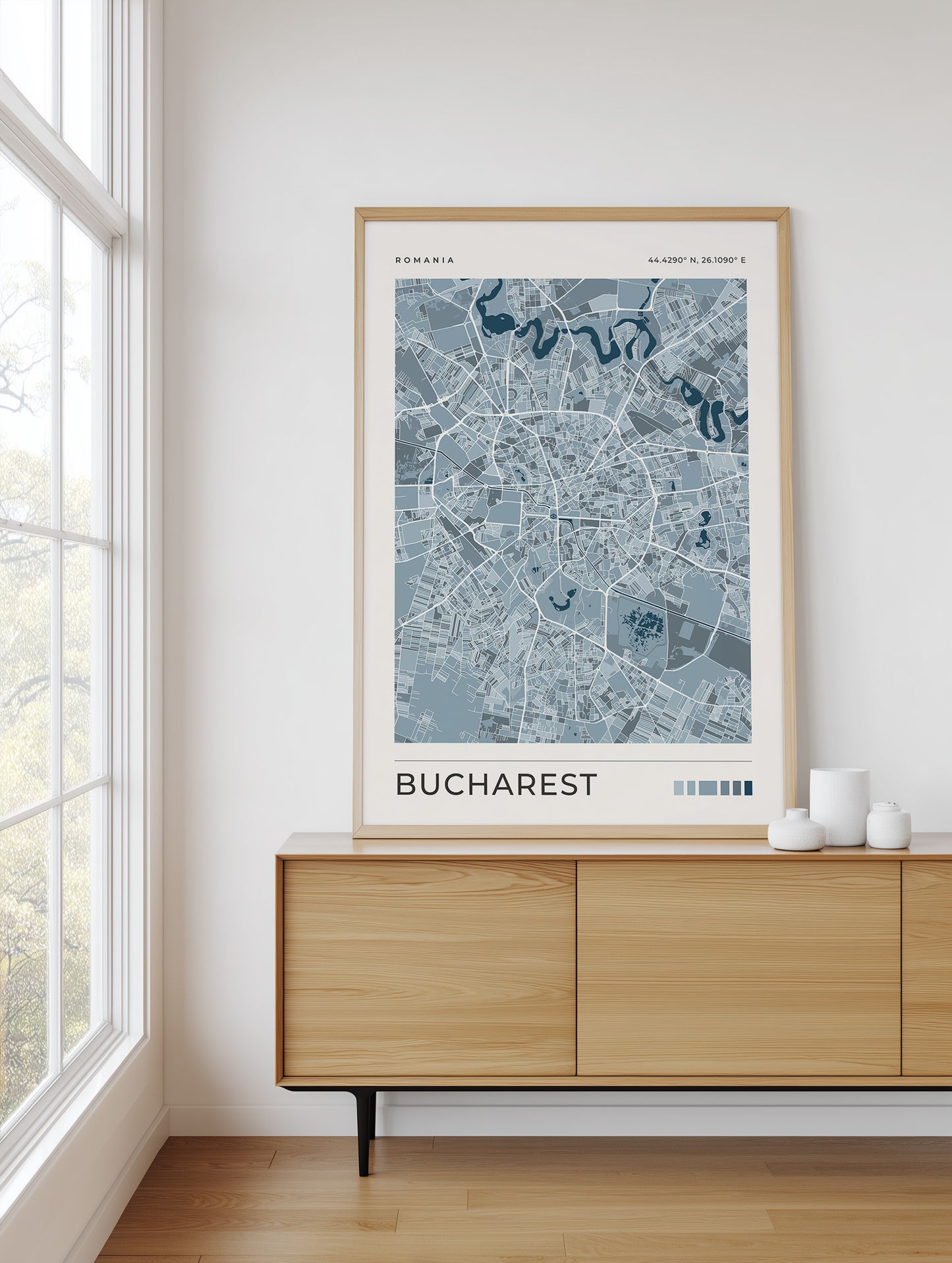 A framed street map art print of Bucharest, Romania, featuring a light blue and gray map design with a light wood frame, displayed on a wooden sideboard in a bright room next to a window.