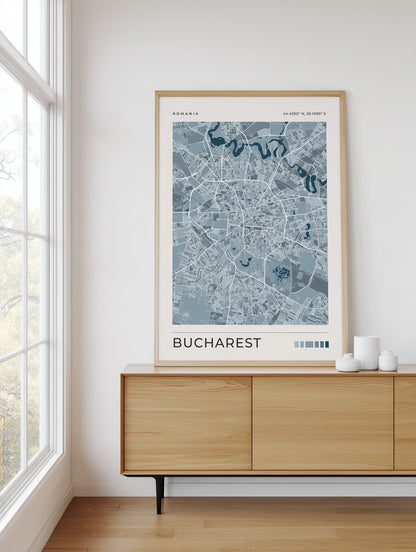 A framed street map art print of Bucharest, Romania, featuring a light blue and gray map design with a light wood frame, displayed on a wooden sideboard in a bright room next to a window.