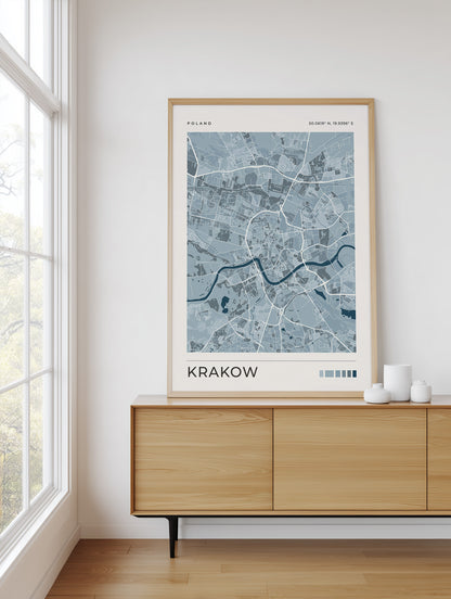 A framed city map print of Krakow, Poland, styled on a light wood sideboard in a bright, modern room next to a window.
