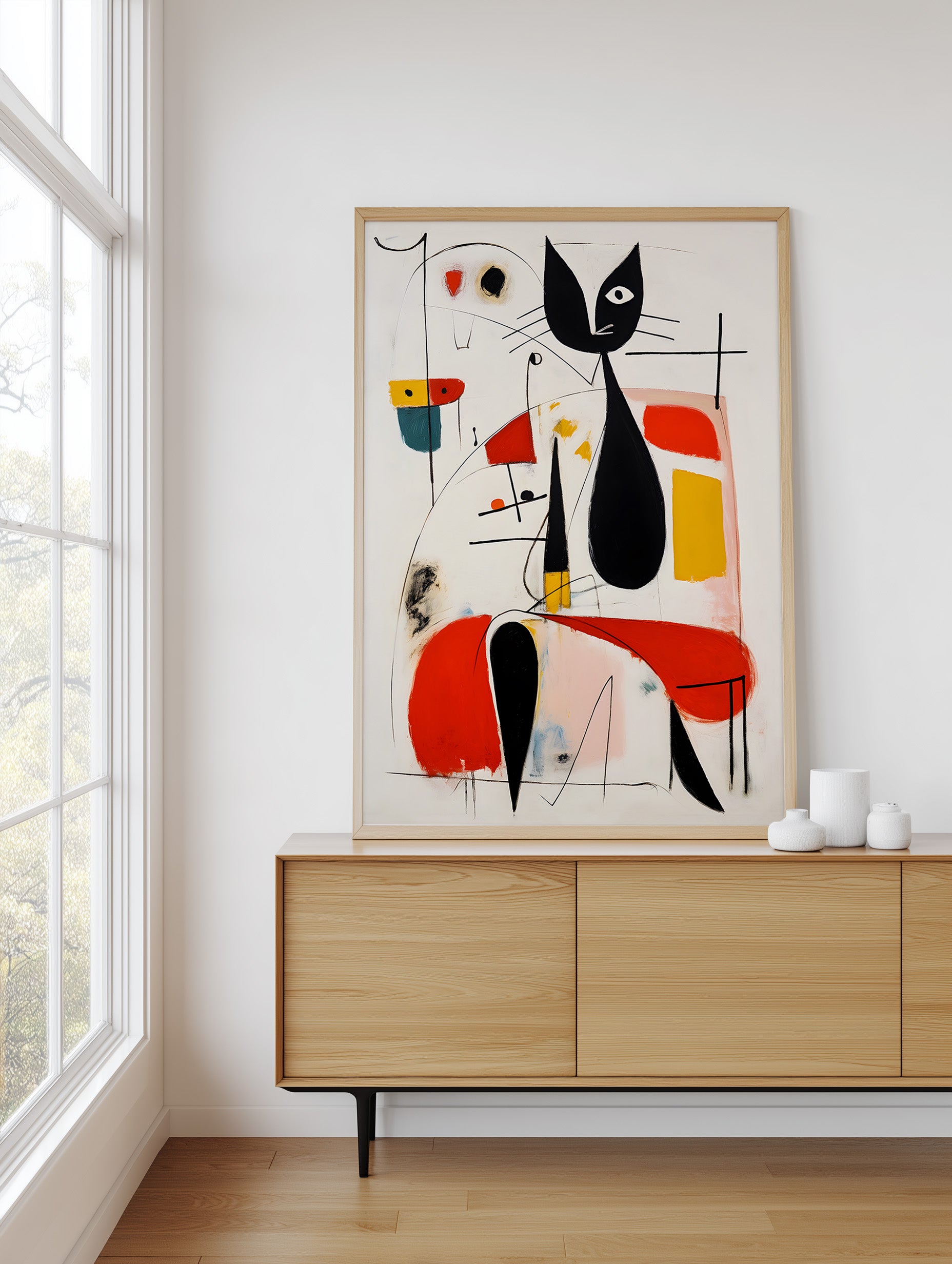 A framed abstract art print featuring a stylized black cat in a minimalist composition, displayed on a light wood sideboard in a bright, modern room next to a window.