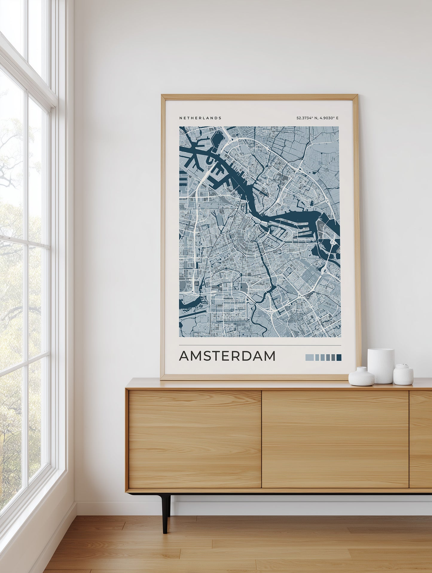 A framed city map print of Amsterdam, Netherlands, displayed on a light wood sideboard in a bright room with a window and white walls.