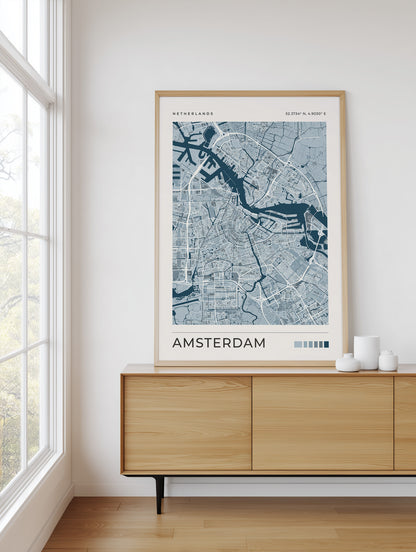 A framed city map print of Amsterdam, Netherlands, displayed on a light wood sideboard in a bright room with a window and white walls.