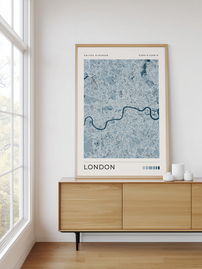 A framed London city map poster in light blue and white tones, displayed on a light wood sideboard in a bright room next to a window.