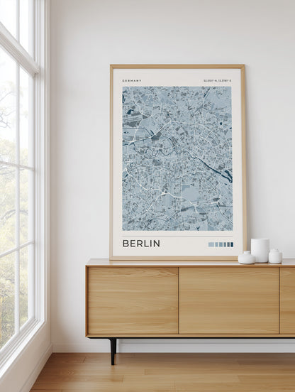 A framed art print of a light blue and white Berlin city map with a light wood frame, displayed on a light wood sideboard in a bright room next to a window.