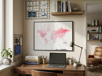 A framed world map poster with pink watercolor continents hanging on a beige wall above a wooden desk in a styled home office setting.
