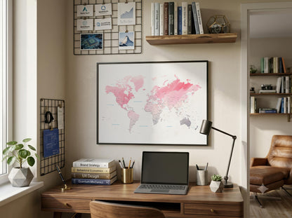 A framed world map print with pink and gray watercolor continents hangs on a beige wall above a wooden desk in a styled home office. The scene includes a laptop, desk lamp, books, plants, shelves, and a chair in the adjacent room.