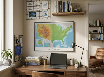 A framed United States topographic map poster hangs on a beige wall above a wooden desk in a styled home office setting with a laptop, lamp, books, and plants.