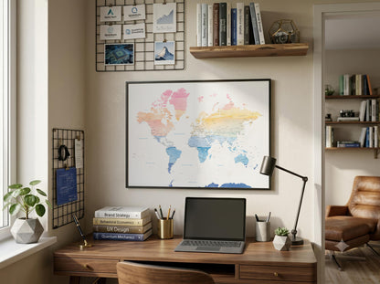 A framed world map poster with pastel-colored continents hangs on a beige wall above a wooden desk in a styled home office setting with a laptop, lamp, and plants.