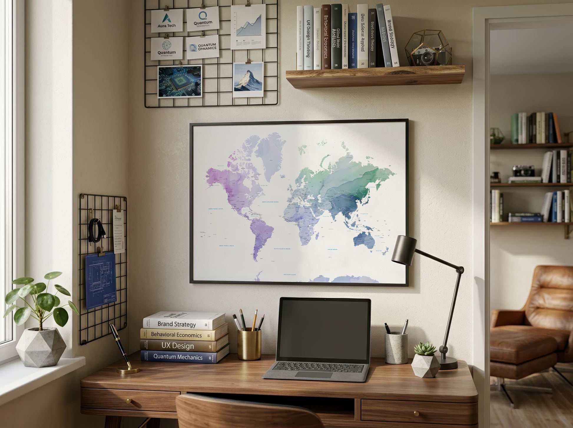 A framed world map poster with a watercolor design hangs on a beige wall above a wooden desk in a styled home office setting. The desk is equipped with a laptop, lamp, books, and plants, with shelves and a wire grid board on the wall.