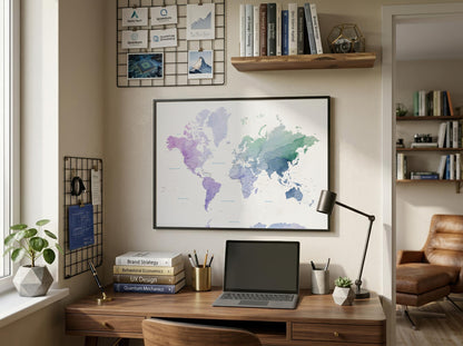 A framed world map poster with a watercolor design hangs on a beige wall above a wooden desk in a styled home office setting. The desk is equipped with a laptop, lamp, books, and plants, with shelves and a wire grid board on the wall.