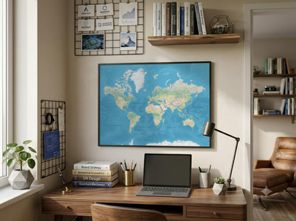 A framed world map with a blue ocean and green/beige landmasses hangs on a beige wall above a wooden desk in a styled home office setting with a laptop, lamp, and shelves.