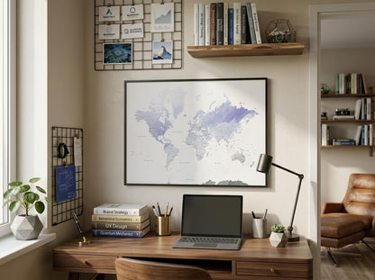 A framed world map print with a soft purple and blue watercolor design hangs on a beige wall above a wooden desk in a styled home office setting.