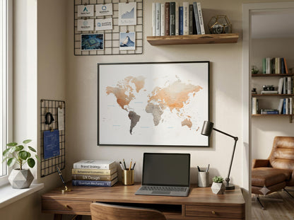 A framed world map poster with a watercolor-style design in muted orange, beige, and gray tones, displayed on a beige wall above a wooden desk in a styled home office setting.