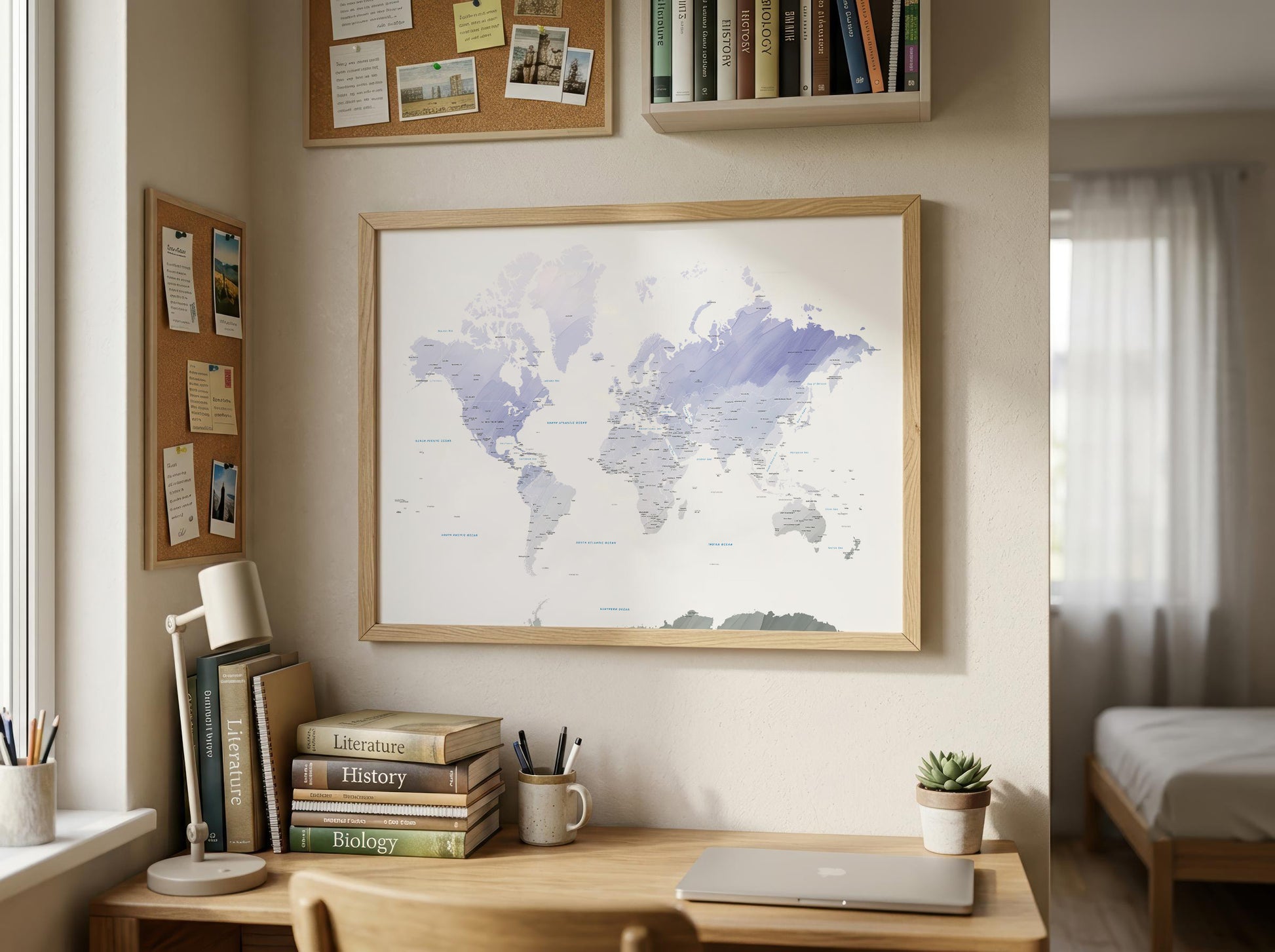 A framed world map print with a light wood frame hangs on a beige wall in a bright, modern room. Below the map is a wooden desk with books, a lamp, a laptop, and a small potted succulent.