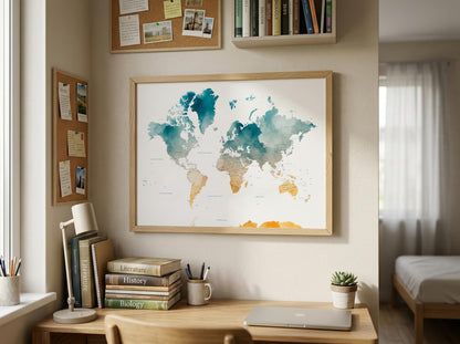 A framed world map poster with a watercolor design hangs on a beige wall in a styled home office or bedroom setting. The room features a wooden desk with books, a lamp, a laptop, and a small succulent, with a window and bed visible in the background.