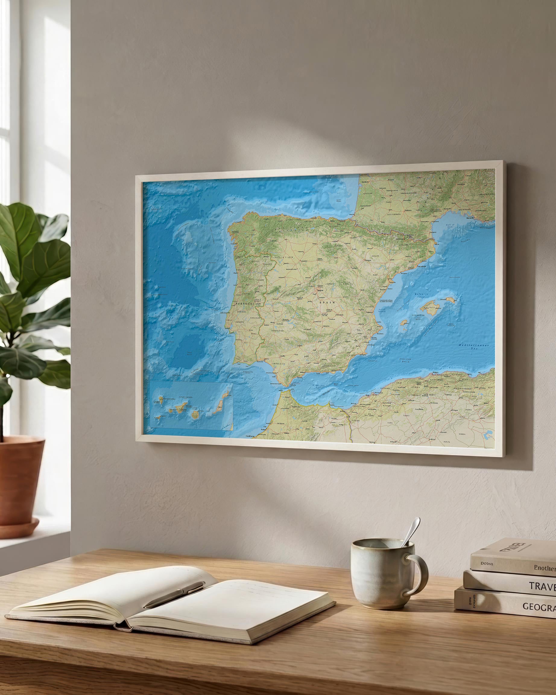 A framed world map print featuring Spain and surrounding Europe, displayed on a beige wall above a wooden desk with an open notebook, mug, and books.