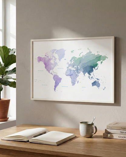 A framed world map poster with a watercolor-style multicolor design in purple, blue, and green tones, hanging on a beige wall above a wooden desk with an open book, a mug, and stacked books.