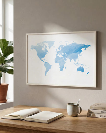 A framed world map art print in shades of blue on a white background, hanging on a beige wall above a wooden desk with an open book, a mug, and stacked books.
