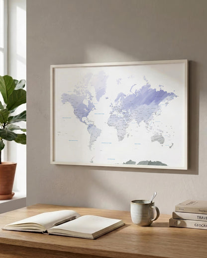 A framed world map print with a white background and light blue watercolor country outlines, hanging on a beige wall above a wooden desk with an open book, a mug, and stacked books.