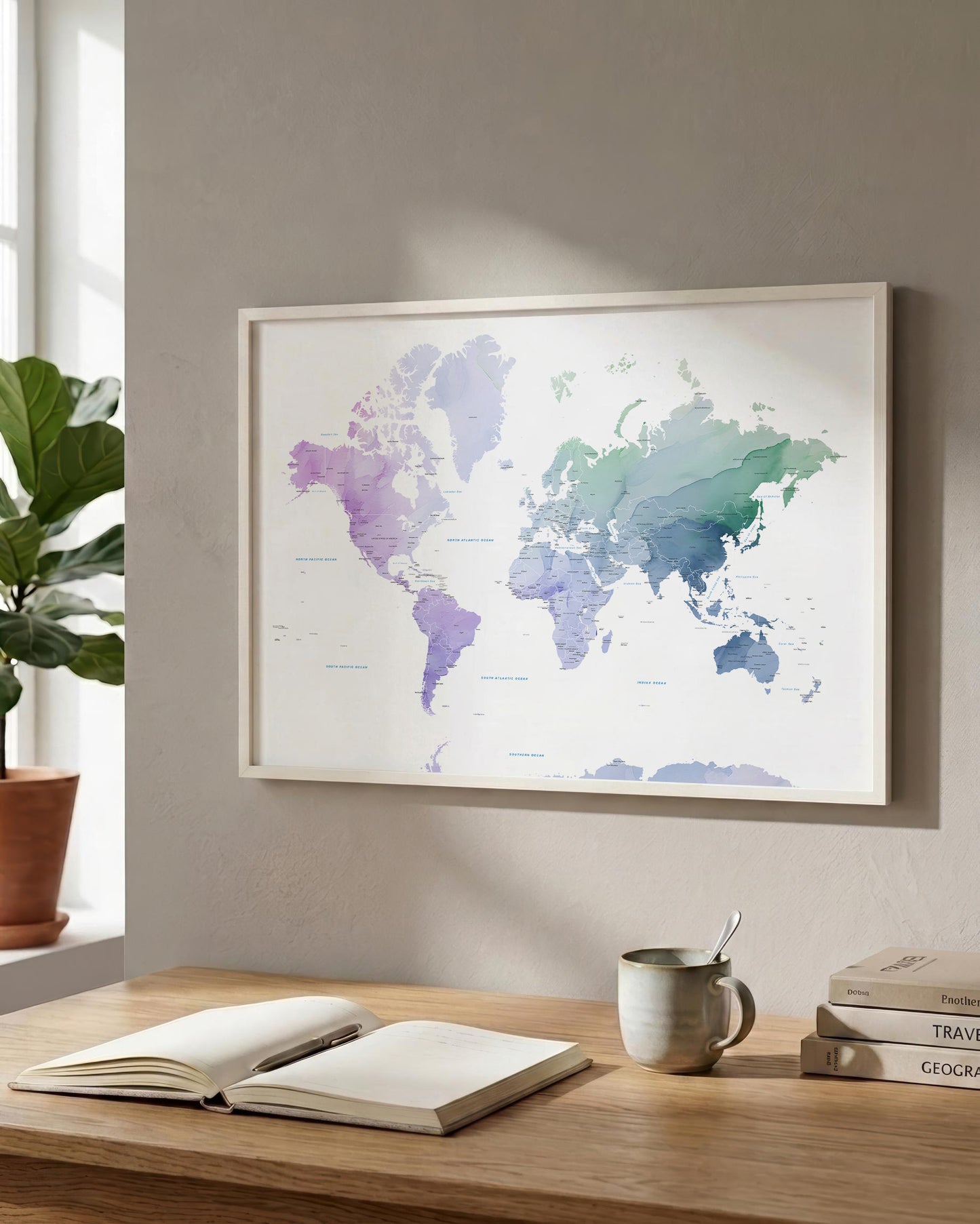A framed world map print with a watercolor design in shades of purple, blue, and green, hanging on a beige wall above a wooden desk with an open book, a mug, and stacked books.