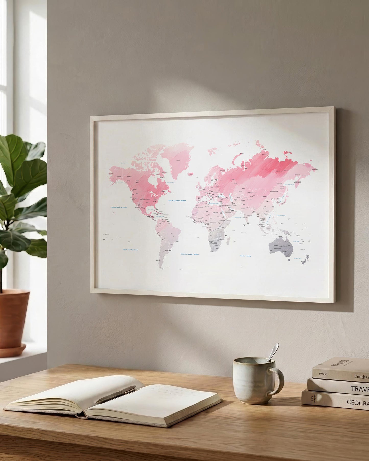 A framed world map print with pink and gray watercolor-style continents hangs on a beige wall above a wooden desk with an open notebook, a mug, and stacked books.