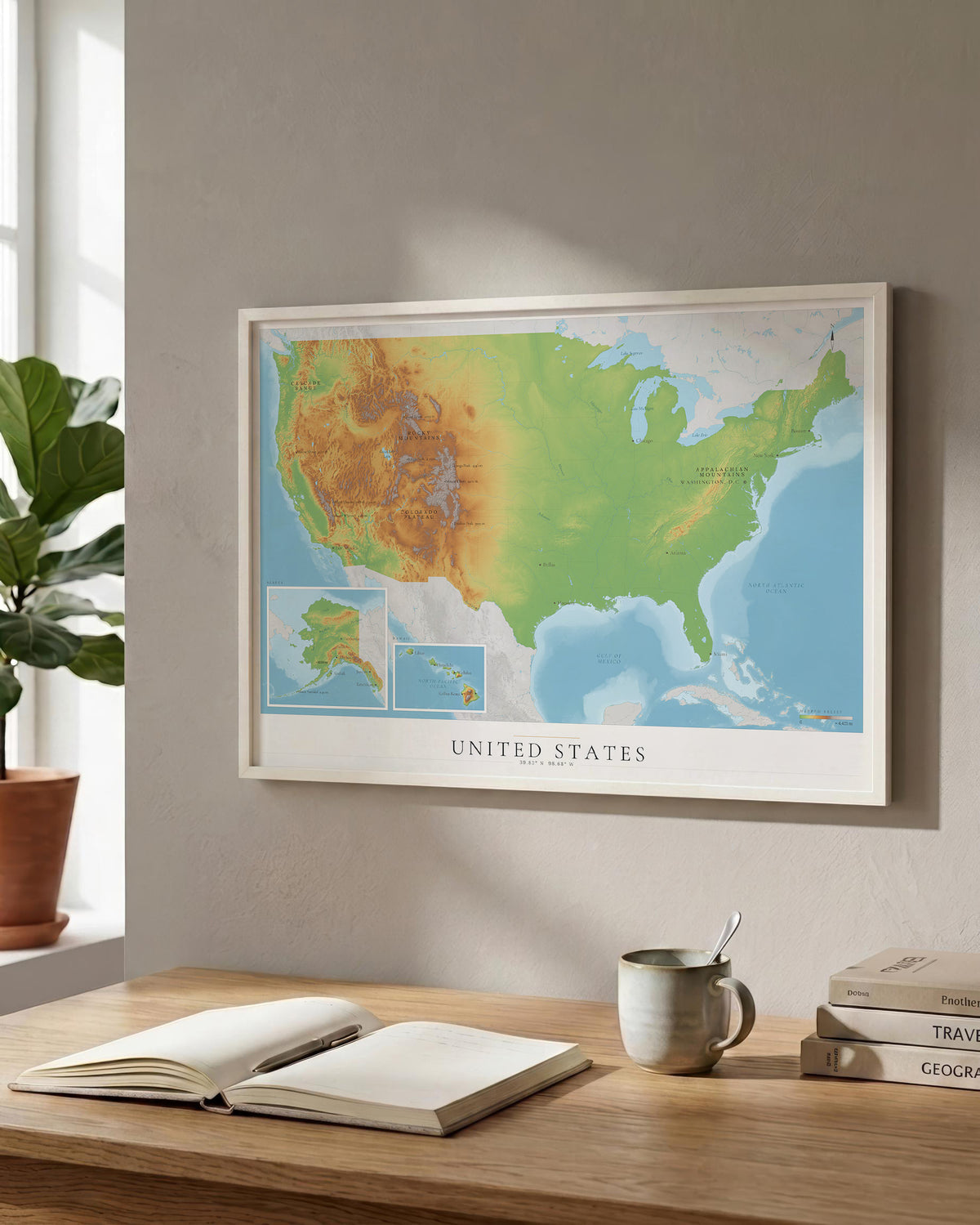 A framed United States topographic map print hangs on a light gray wall above a wooden desk with an open book, a ceramic mug, and stacked books. A potted plant is visible at the window on the left.