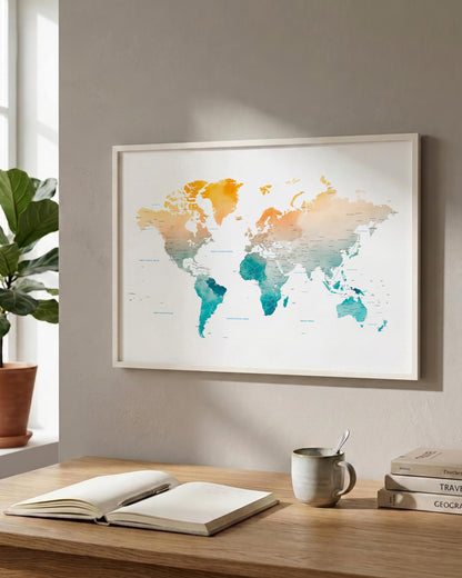 A framed world map print with a watercolor finish hangs on a light gray wall above a wooden desk. The map features warm orange and yellow landmasses transitioning into teal and blue oceans, set in a clean, modern room with a potted plant, an open notebook, and a ceramic mug.