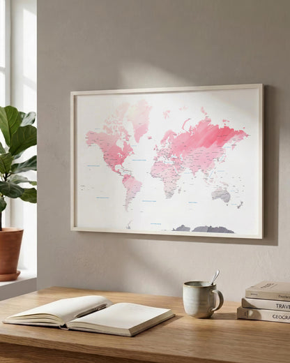 A framed world map print with pink watercolor country outlines hangs on a light gray wall above a wooden desk with an open book, a mug, and stacked books. A potted plant is visible at the window on the left.